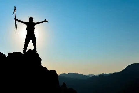 Silhouette einer Person mit Wanderstöcken auf einem Berggipfel, die ihre Arme ausbreitet. Ein Symbol für das Erreichen von Zielen im Leben.