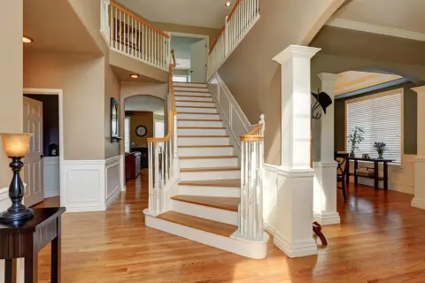 A grand foyer with a sweeping staircase featuring white painted risers and wooden treads. The staircase leads to an upper landing, with a view into a living area.