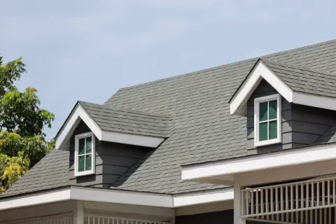 A close-up of a house roof showcasing different types of shingles in a grey hue, with dormer windows peeking out.