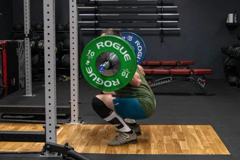 Mężczyzna wykonuje głęboki **squat** z ciężarem na plecach w siłowni. Zielone talerze Rogue ważą 10 kg każdy.
