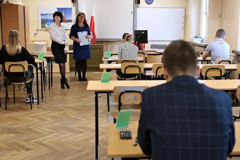 Uczniowie siedzą w ławkach, czekając na egzamin. Nauczycielki stoją z przodu, trzymając dokumenty. Ile trzeba mieć procent żeby zdać egzamin zawodowy?