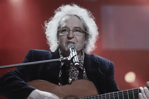 Zegarmistrz światła purpurowy, z burzą białych włosów, gra na gitarze i śpiewa przy mikrofonie.