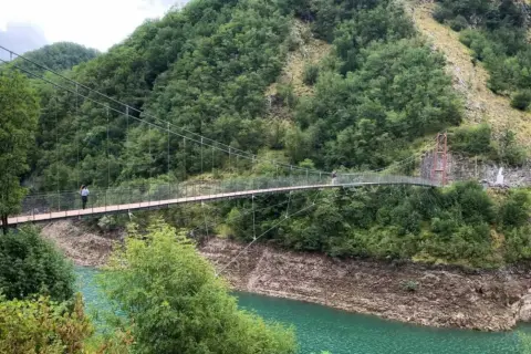 Un ponte sospeso attraversa un lago turchese, circondato da montagne boscose. Due persone passeggiano sul ponte, godendosi il panorama dei vagli di sotto.