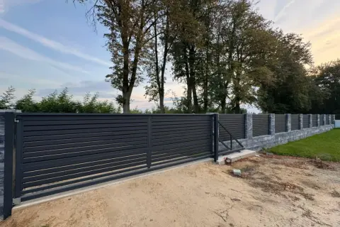 Nowoczesne, ciemnoszare ogrodzenie z przesuwną bramą. Widać kamienny murek i drzewa w tle. Odległość ogrodzenia od drogi gminnej wewnętrznej jest niewielka.