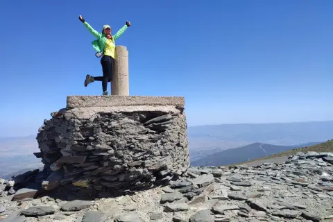 Mujer con el chullo puesto, brazos en alto, celebra en la cima de una montaña rocosa bajo un cielo azul despejado.