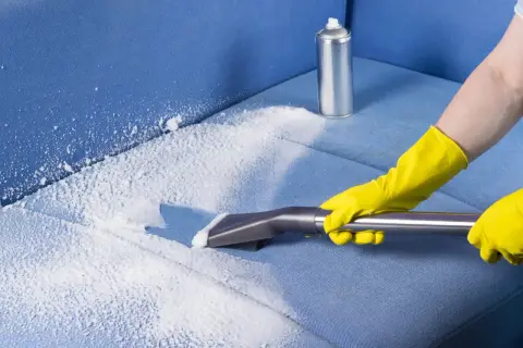 Mani con guanti gialli eseguono il lavaggio divano a secco con schiuma e aspiratore su tessuto blu.