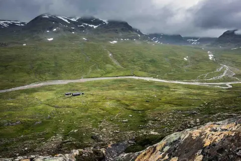 Grönskande fjällandskap med en slingrande älv och små stugor. En del av Kungsleden vid Hemavan, med snö på topparna.