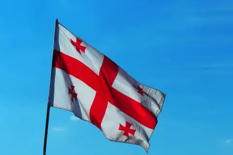 Flaga Gruzji powiewa na tle błękitnego nieba. Biały materiał ozdobiony jest dużym czerwonym krzyżem i czterema mniejszymi krzyżami w rogach.