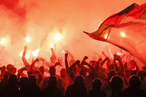 Tłum kibiców w czerwonym dymie z rac, z uniesionymi pięściami i flagą. Widać też sylwetki policjantów w hełmach.