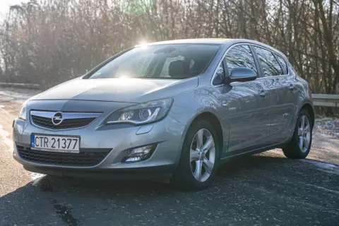 Szary Opel Astra 2011 na drodze, z drzewami w tle i słońcem odbijającym się od karoserii.
