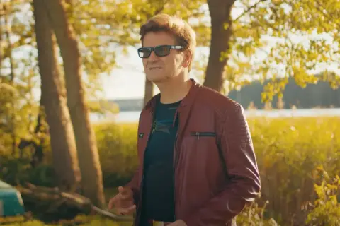 Mężczyzna w skórzanej kurtce, niczym król imperium disco polo, pozuje na tle jesiennego krajobrazu nad jeziorem.