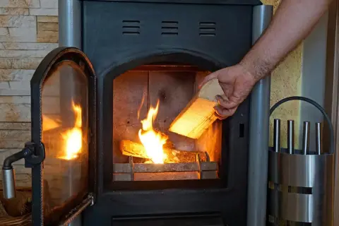 Hand legt Holzscheit in brennenden Kaminofen. Beachten Sie die Kaminofen Verordnung für sicheres Heizen.