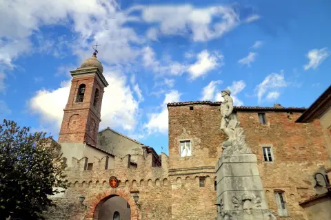 Poggio San Marcello: campanile in mattoni, un arco fortificato e una statua di bronzo sotto un cielo azzurro con nuvole bianche.