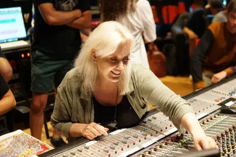 Sylvia Massy, lächelnd, bedient ein Mischpult in einem Tonstudio. Menschen im Hintergrund sind beschäftigt.