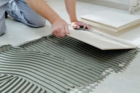 Hands carefully place a tile over cement board, with a stack of tiles nearby.