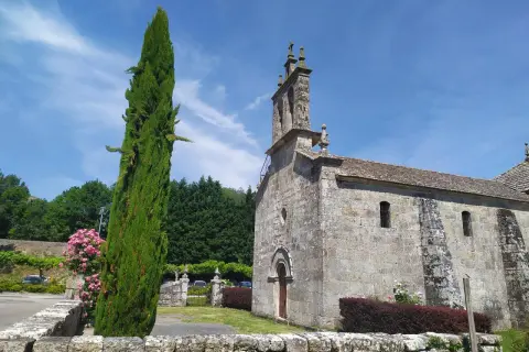 Iglesia de piedra bajo cielo azul, con un ciprés alto y flores rosas. Un lugar tranquilo en Cova da Moura.