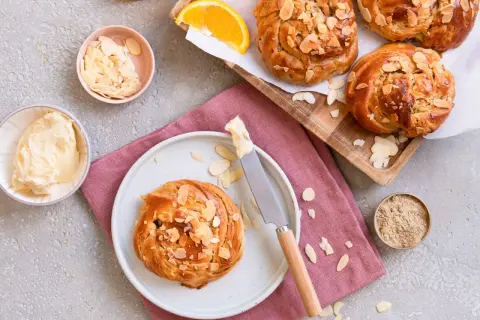 Goldbraune Kardamom-Knoten, bestreut mit Mandelblättchen, warten auf Butter und Orangenscheiben.