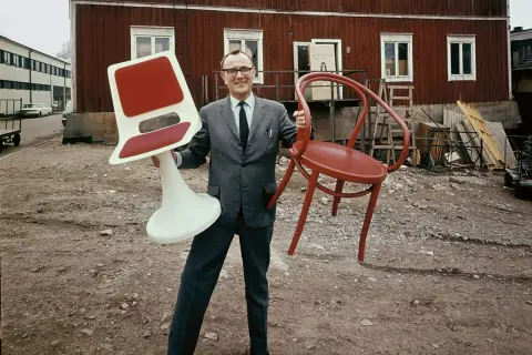 Man i kostym håller en röd stol och en vit stol med röd sits. Kanske en tidig idé för Ingvar Kamprad utbildning i möbeldesign.
