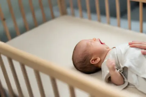 A baby yawns in a wooden crib. This image reassures parents that mini cribs are safe for their little ones.