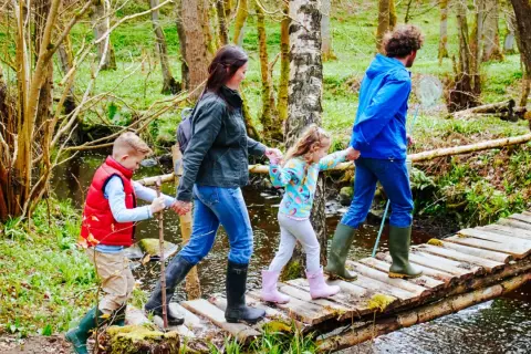Familie überquert Holzbrücke auf einem erlebnispfad nrw. Vater hält Kescher, Kinder halten sich an Eltern.