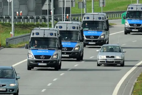 Konwój pojazdów uprzywilejowanych: policyjne furgonetki i samochód osobowy jadące autostradą.