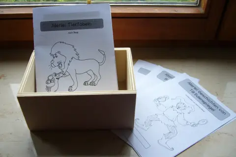 Holzkiste mit Blättern für fabeln zum lesen. Ein Blatt zeigt einen Löwen mit einem Lamm, ein anderes einen Till Eulenspiegel.