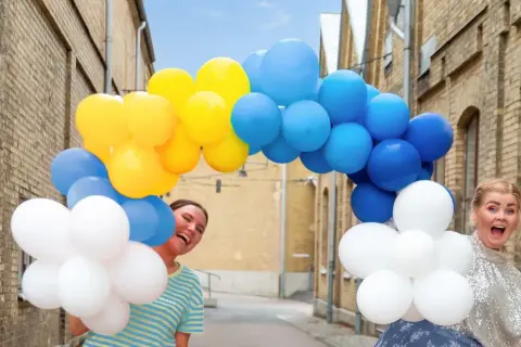 Två glada kvinnor firar en studentfest med en båge av blå, gula och vita ballonger.