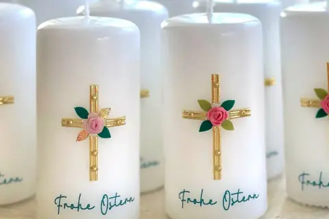 Mehrere weiße Osterkerzen mit goldenen Kreuzen, rosa Rosen und grünen Blättern. Die Osterkerze Bedeutung wird durch diese Symbole unterstrichen. "Frohe Ostern" steht geschrieben.