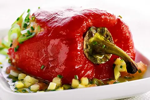 En fylld, röd paprika serveras på en vit tallrik med en sallad av tärnad paprika och örter. Ett läckert recept med grillad paprika på burk.