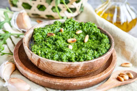 Frisches Rucola Pesto in Holzschale, garniert mit Pinienkernen. Daneben Knoblauch und Olivenöl.