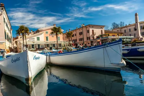 Barche a Cisano di Bardolino, con palme e case colorate sullo sfondo. Il sole splende su un cielo azzurro e limpido.