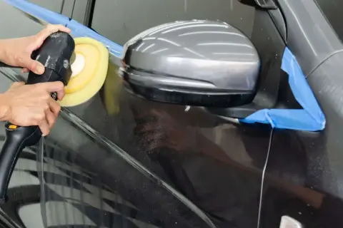 Ręce trzymają polerkę, która wykonuje polerowanie lakieru bezbarwnego na ciemnym samochodzie.