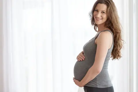 Uśmiechnięta ciężarna kobieta trzymająca się za brzuch, czując delikatne ciągnięcie brzucha w ciąży podczas chodzenia.