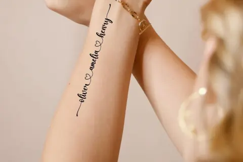 Ein zartes **Enkel Tattoo Namen** zeigt "oliver & amelia & henry" in geschwungener Schrift auf einem Arm, geschmückt mit einem goldenen Armband.