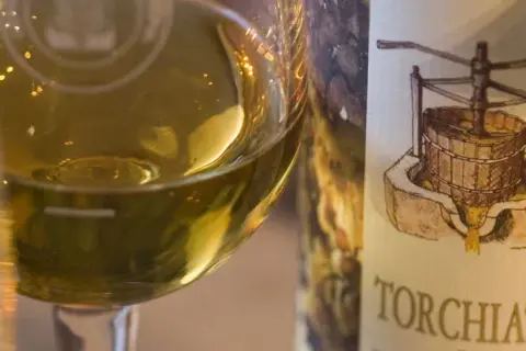 Calice di vino bianco dorato accanto a una bottiglia con etichetta che raffigura un torchio e grappoli d'uva, evocando il sapore del torchiato di fregona.