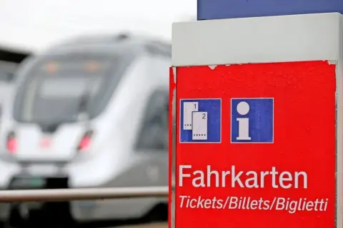 Roter Fahrkartenautomat am Bahnhof. Ein Zug fährt vorbei. Wann am besten Bahntickets buchen? Hier gibt es Infos.
