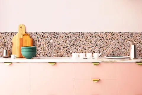 Cuisine moderne avec des armoires roses, un plan de travail en marbre et un crédence en terrazzo coloré. Ustensiles de cuisine et vaisselle disposés.