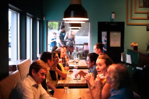 Ludzie siedzą przy stołach w przytulnym bistro, rozmawiają i piją.
