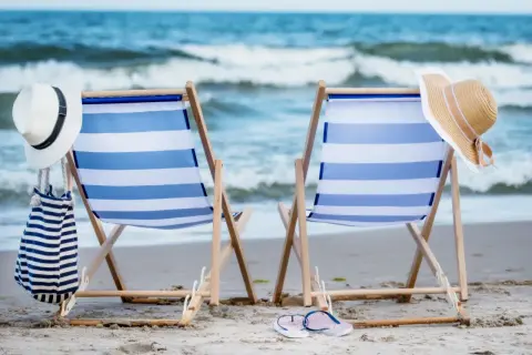 Dwa leżaki plażowe z niebiesko-białymi pasami, kapelusze i torba na piasku, z widokiem na morze.