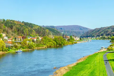 Blick auf den Elberadweg mit Radfahrern und einer Stadt am Flussufer. Die Elbe fließt durch eine grüne Landschaft mit bewaldeten Hügeln. Die Elberadweg Entfernungen sind hier gut zu erkunden.