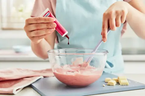 En hand droppar röd färg i en skål med rosa smet, för att skapa ett perfekt glasyr recept.