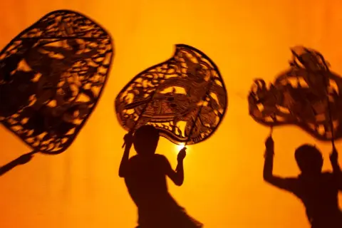 Trzy postacie z papierowymi lalkami tworzą cienie na pomarańczowym tle. Tak właśnie na czym polega teatr cieni – magia światła i wyobraźni.