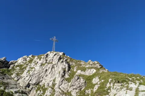 Na szczycie skalistego zbocza, porośniętego trawą, stoi metalowy krzyż. W tle błękitne niebo, a w oddali widać samolot. To widok z Giewontu, który przypomina, ile ma wysokości.