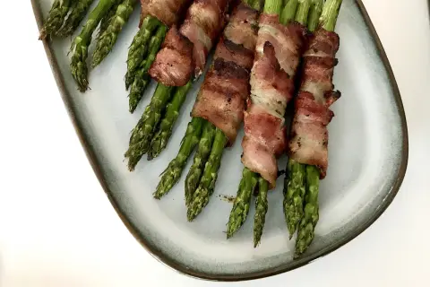 Grön sparris insvept i knaprigt bacon, serverad på en ljusblå tallrik. En utsökt kombination av grönsaker och baconlindad sparris.