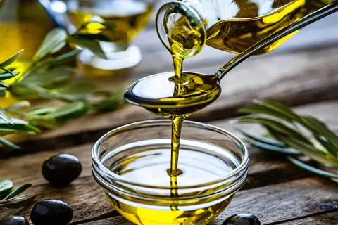 Olio di oliva non extravergine versato da un cucchiaio in una ciotola, con olive e rami d'ulivo sullo sfondo.