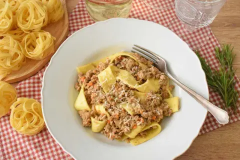 Pappardelle z bogatym ragu, które to danie, posypane parmezanem. Obok surowy makaron i gałązka rozmarynu.