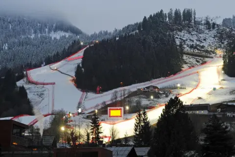 Die Skirennstrecke, beleuchtet für die Nacht, mit roten Banden und einem roten Hahnkamm-Symbol.