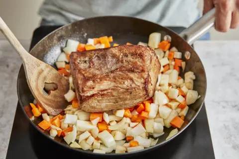Ein gebratener Rinderbraten ruht auf gewürfeltem Wurzelgemüse in einer Pfanne. Perfekt für ein herzhaftes rinderbraten rezept.