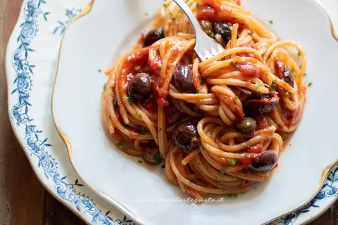 Un piatto invitante di spaghetti alla puttanesca, con olive nere, capperi e un sugo rosso vivace, pronto per essere gustato.