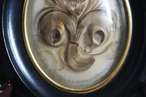 Intricate hair art in a framed oval, featuring swirling locks of blonde hair arranged like a floral bouquet. Text reads "A Marie, une mère la remercie et lui demande de protéger son fils Jean Boudet.